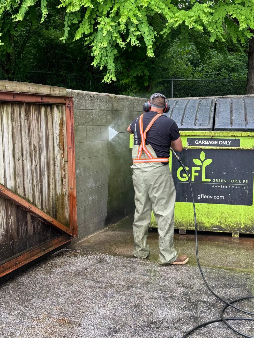 A Deoro crew member in high-visibility braces pressure-washing the block wall of a dumpster enclosure.