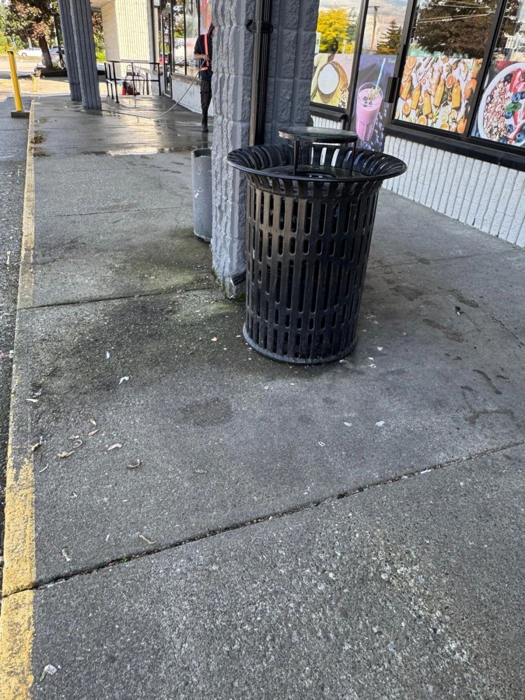 A storefront walkway beside a waste bin, the concrete darkly stained and scattered with debris, a crew member pressure-washing in the background.
